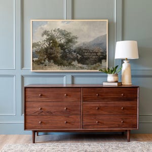 May include: A wooden dresser with six drawers, a framed landscape painting, and a table lamp. The painting depicts a tree and mountains with text. The lamp has a white shade and a cream-colored base. A small potted plant sits on a stack of books.