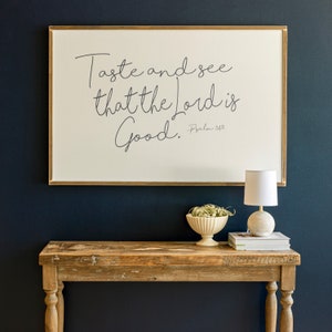May include: A framed print with the text "Taste and see that the Lord is good. Psalm 34:8" hanging on a dark blue wall above a wooden console table with a small lamp and a bowl of greenery.