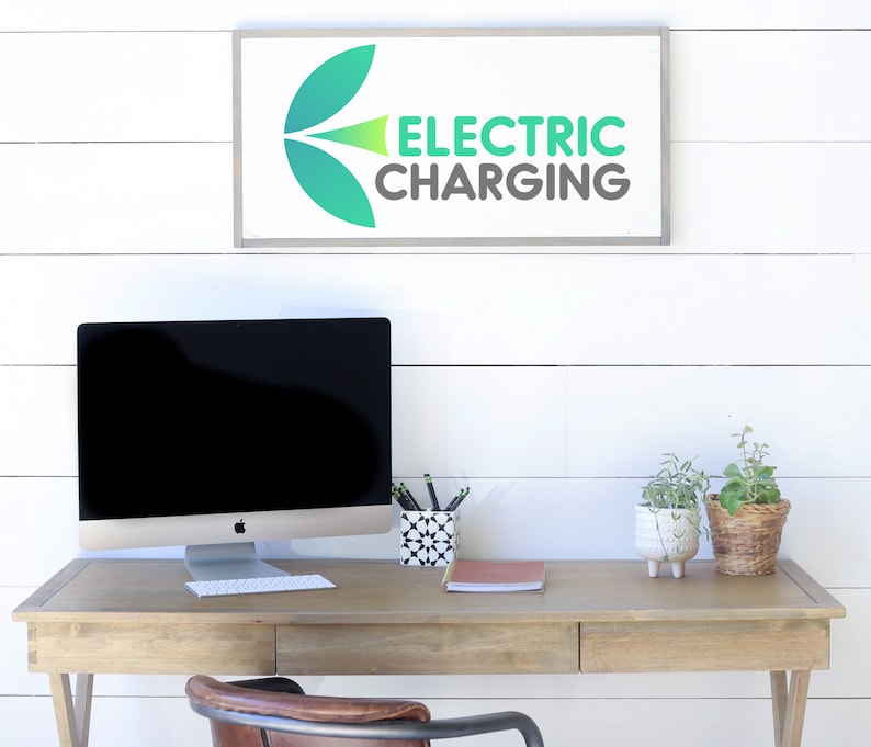 May include: A wooden desk with a computer, keyboard, and accessories. A sign above the desk reads "ELECTRIC CHARGING" in green and gray. Two small potted plants and a pen holder are on the desk.