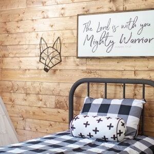 May include: A black metal bed frame with a white and black checkered bedding set. A white pillow with black plus signs is on the bed. A wooden wall with a black metal fox sculpture and a white sign with the text "The Lord is with you Mighty Warrior" is behind the bed.