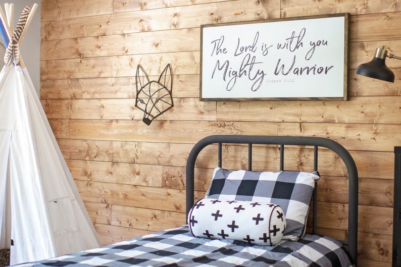 May include: A black metal bed frame with a white and black checkered bedding set. A white pillow with black plus signs is on the bed. A wooden wall with a black metal fox sculpture and a white sign with the text "The Lord is with you Mighty Warrior" is behind the bed.