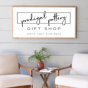 May include: A framed sign with the text "prodigal pottery GIFT SHOP GIFTS THAT GIVE BACK" hangs above two cream-colored armchairs. A small wooden table with a vase and greenery sits between the chairs. The wall is painted light gray.