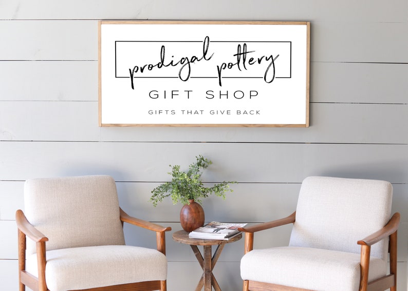 May include: A framed sign with the text "prodigal pottery GIFT SHOP GIFTS THAT GIVE BACK" hangs above two cream-colored armchairs. A small wooden table with a vase and greenery sits between the chairs. The wall is painted light gray.