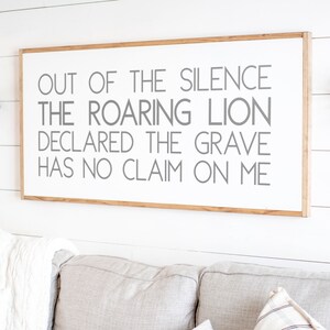 Może przedstawiać: Biały drewniany znak z drewnianą ramą wisi na białej ścianie. Znak zawiera tekst "OUT OF THE SILENCE THE ROARING LION DECLARED THE GRAVE HAS NO CLAIM ON ME" szarymi literami.
