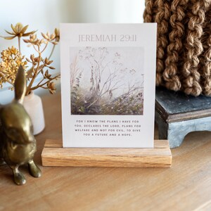 May include: A card with the text "JEREMIAH 29:11" and a quote about plans and hope, displayed in a wooden holder. The card features a watercolor image of a field. A brass bunny figurine and dried flowers are in the background.