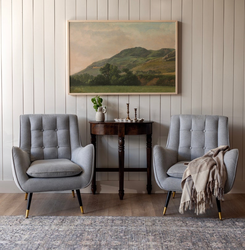 Op de afbeelding: Twee grijze gestoffeerde fauteuils met knoopbekleding, een houten tafel met een dienblad en kandelaars, en een ingelijst landschapsschilderij op een witte muur.