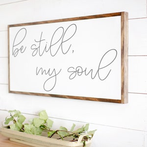 May include: A white wooden sign with a brown wood frame, hanging on a white wall. The sign has the text "Be still, my soul" written in cursive gray lettering.