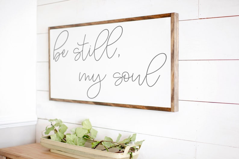 May include: A white wooden sign with a brown wood frame, hanging on a white wall. The sign has the text "Be still, my soul" written in cursive gray lettering.