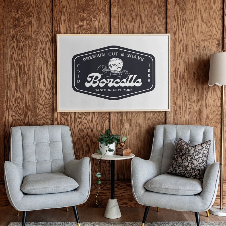 May include: A framed sign with the text "Premium Cut & Shave" and "Borcello Based in New York" hangs on a wood-paneled wall. Two gray armchairs with button-tufted backs and black and gold legs face the sign. A small side table with a marble top and a floor lamp complete the scene.