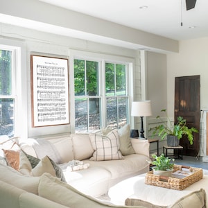 May include: A white sectional sofa with multiple pillows in a living room setting. The sofa is positioned in front of a large window with a view of trees. A framed print of sheet music with the title "How Great Thou Art" hangs on the wall above the sofa.