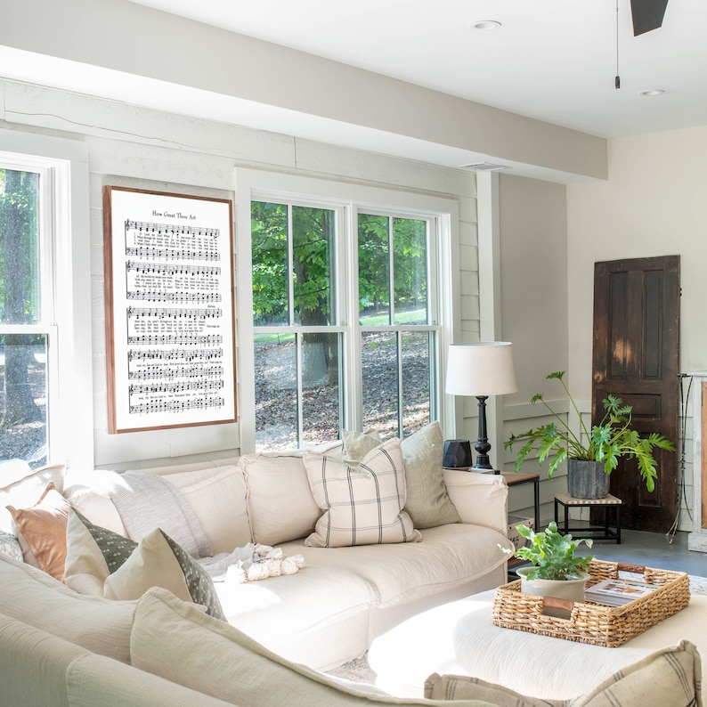 May include: A white sectional sofa with multiple pillows in a living room setting. The sofa is positioned in front of a large window with a view of trees. A framed print of sheet music with the title "How Great Thou Art" hangs on the wall above the sofa.