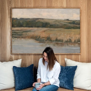 May include: A rectangular landscape painting in a light wood frame featuring a muted palette of beige, brown, and green tones depicting a grassy field near a body of water under a cloudy sky, mounted on a vertical light wood paneled wall.