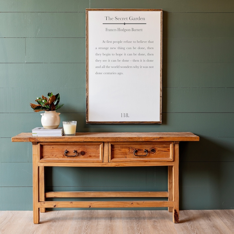 May include: A wooden console table with two drawers and a framed print with a quote from "The Secret Garden" by Frances Hodgson Burnett. The quote reads: "At first people refuse to believe that a strange new thing can be done, then they begin to hope it can be done, then they see it can be done - then it is done and all the world wonders why it was not done centuries ago." The number 118 is printed below the quote.