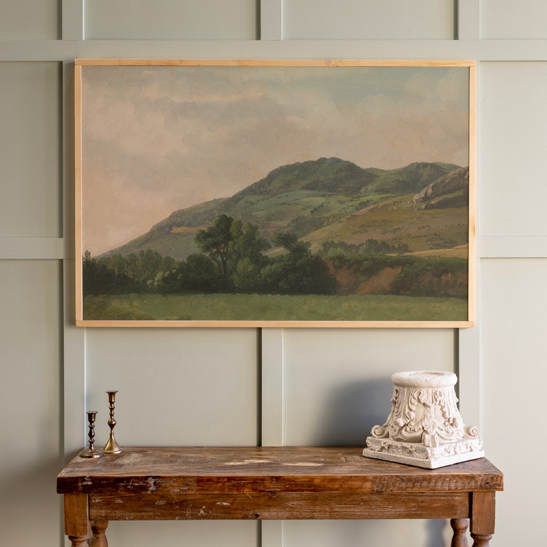 Op de afbeelding: Een ingelijst landschapschilderij dat een glooiende groene heuvel met een bewolkte lucht op de achtergrond toont. Het schilderij is in een houten lijst en hangt aan een lichtgroene muur.
