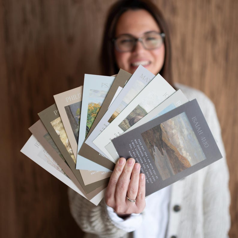 May include: A collection of cards with landscape art and text, fanned out in a hand. The cards have a variety of colors, including beige, gray, and blue. One card features the text "ISAIAH 41:10".