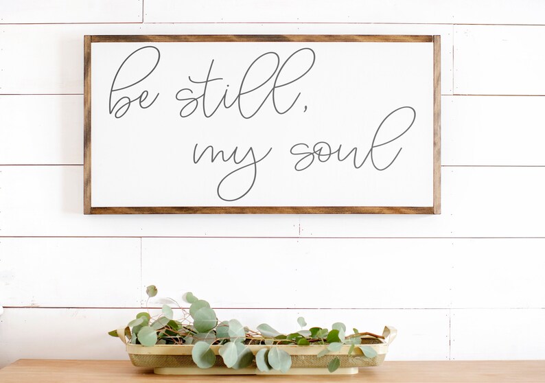 May include: A white wooden sign with a brown frame, featuring the text "be still, my soul" in cursive script.