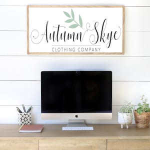 May include: A wooden desk with a computer, keyboard, and office supplies. Above the desk is a framed sign that reads "Autumn Skye Clothing Company" in elegant script. The sign has a light wood frame and a white background.