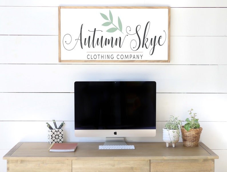 May include: A wooden desk with a computer, keyboard, and office supplies. Above the desk is a framed sign that reads "Autumn Skye Clothing Company" in elegant script. The sign has a light wood frame and a white background.