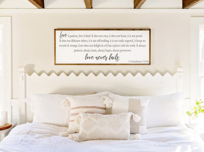Op de afbeelding: Een wit houten bord met een bruin frame hangt boven een wit bed met een wit hoofdeinde. Het bord heeft een grijze inscriptie die luidt: "Liefde is geduldig, liefde is vriendelijk. Ze is niet jaloers, ze is niet opschepperig, ze is niet trots. Ze handelt niet onfatsoenlijk, ze zoekt niet haar eigen belang, ze is niet snel boos, ze houdt geen rekening met kwaad. Liefde verheugt zich niet over onrecht, maar verheugt zich over de waarheid. Ze beschermt altijd, ze vertrouwt altijd, ze hoopt altijd, ze volhardt altijd. Liefde faalt nooit. 1 Korinti&euml;rs 13:4-8"