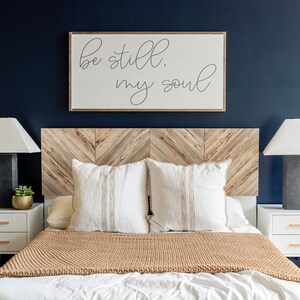 May include: A bedroom with a wooden headboard featuring a chevron pattern. The headboard is decorated with a framed print that reads "Be still, my soul". The bed is made with white bedding and a brown knitted throw blanket. Two white pillows with brown stripes are on the bed. Two bedside tables with white drawers are on either side of the bed. Each table has a lamp with a white shade and a small potted plant.