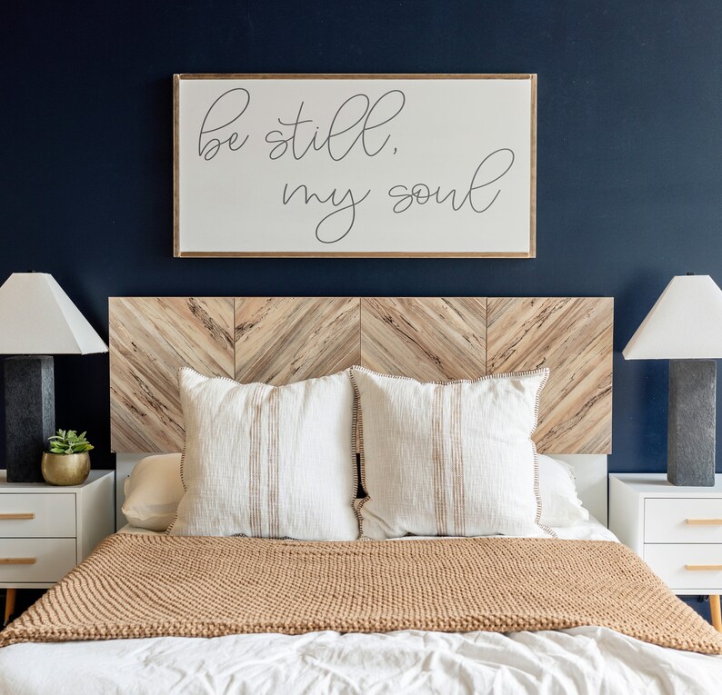 May include: A bedroom with a wooden headboard featuring a chevron pattern. The headboard is decorated with a framed print that reads "Be still, my soul". The bed is made with white bedding and a brown knitted throw blanket. Two white pillows with brown stripes are on the bed. Two bedside tables with white drawers are on either side of the bed. Each table has a lamp with a white shade and a small potted plant.