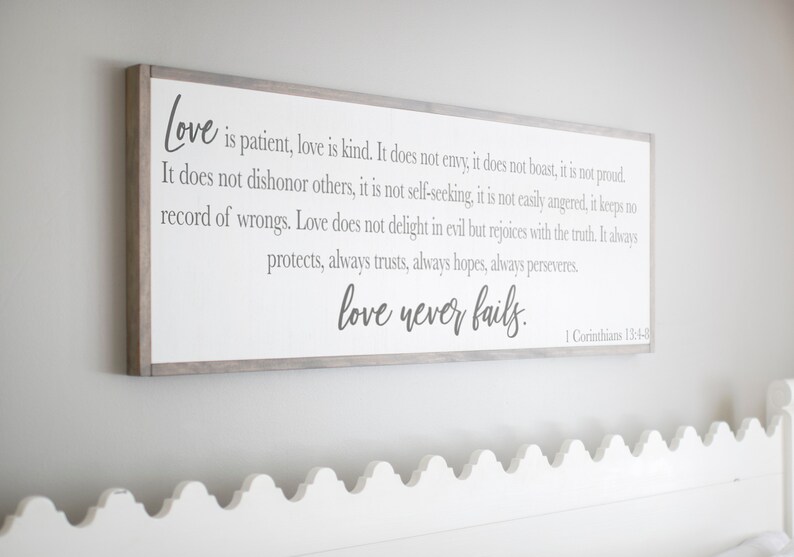 Op de afbeelding: Een ingelijst houten bord met een witte achtergrond en grijze tekst. Het bord bevat een citaat uit de Bijbel, 1 Korinti&euml;rs 13:4-8, over liefde. Het citaat luidt: "De liefde is geduldig, de liefde is vriendelijk. Zij is niet jaloers, zij is niet opschepperig, zij is niet hoogmoedig. Zij is niet onbeleefd, zij zoekt niet haar eigen belang, zij wordt niet verbitterd, zij rekent het kwaad niet aan. De liefde verheugt zich niet over onrecht, maar verheugt zich over de waarheid. Zij verdraagt alles, zij gelooft alles, zij hoopt alles, zij verdraagt alles. De liefde zal nooit ophouden."