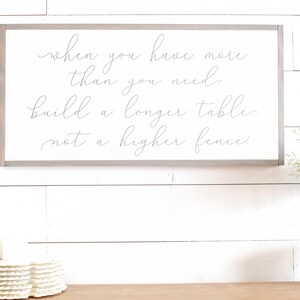 May include: A white wooden sign with a grey frame, featuring the quote "when you have more than you need, build a longer table, not a higher fence."