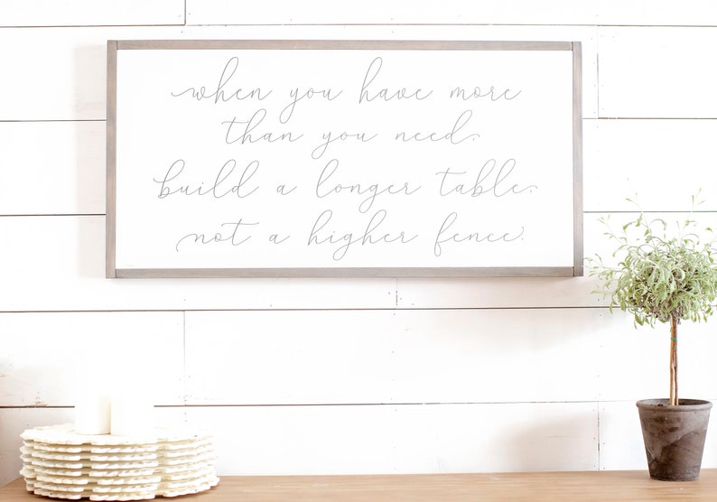 May include: A white wooden sign with a grey frame, featuring the quote "when you have more than you need, build a longer table, not a higher fence."