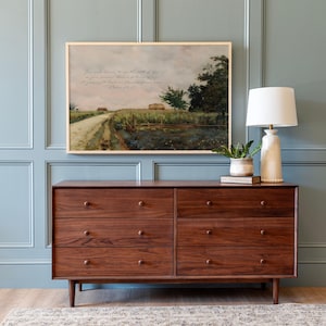 May include: A dark brown six-drawer dresser with a mid-century modern design. The dresser has round knobs and tapered legs. Above the dresser is a framed landscape painting with text. A white lamp and small potted plant sit on top of the dresser.
