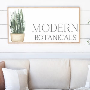 May include: A framed wall art piece with the words "MODERN BOTANICALS" in black lettering. The art features a watercolor illustration of a green snake plant in a beige pot. The frame is a light brown color.