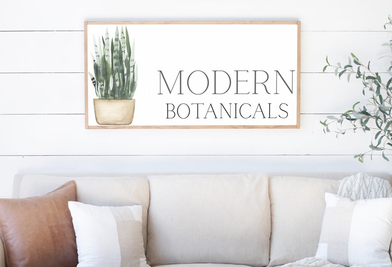 May include: A framed wall art piece with the words "MODERN BOTANICALS" in black lettering. The art features a watercolor illustration of a green snake plant in a beige pot. The frame is a light brown color.