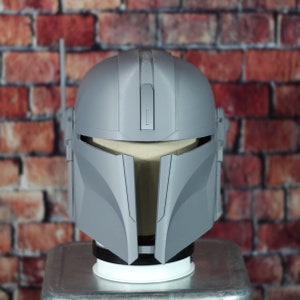 May include: A gray plastic Mandalorian helmet with a visor and a textured surface. The helmet is on a display stand.