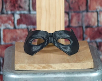 May include: A black eye mask with a textured surface, resting on a wooden block. The mask has eye openings and a curved shape, suggesting a superhero or costume theme. The background features a brick wall.