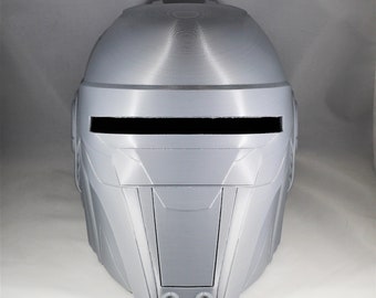 May include: A light gray helmet with a rectangular visor opening and two circular details at the chin. The helmet has a futuristic design with angular panels and a smooth, matte finish. The helmet is likely a prop or costume piece.