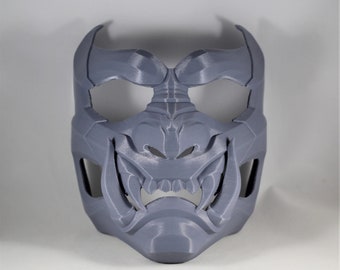 Può includere: Una maschera grigia stampata in 3D con un design dettagliato che ricorda un demone giapponese oni. La maschera ha grandi fori per gli occhi aperti e un'espressione feroce con denti affilati.