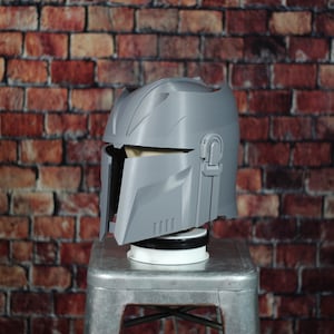 May include: A grey plastic helmet with a textured surface and a visor. The helmet has a design that resembles a Mandalorian helmet.
