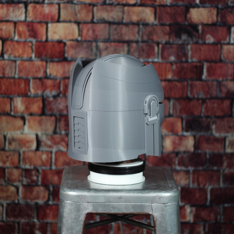 May include: A grey 3D printed helmet with a textured surface and a raised detail on the side. The helmet is sitting on a white and black circular base.