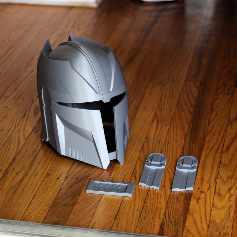 May include: A grey plastic helmet with a visor and a textured surface. The helmet is sitting on a wooden surface with three grey plastic pieces.