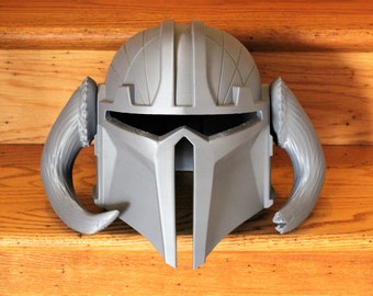 May include: A gray Mandalorian helmet with a distinctive T-shaped visor and curved, horn-like attachments on each side. The helmet features detailed paneling and a smooth, metallic finish, suggesting a futuristic or sci-fi theme.
