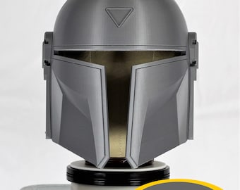 May include: A gray 3D printed helmet with a triangular design on the top. The helmet is on a white base with a black ring around it. The base is on a metal surface. The text "MMCC Approved" is on a yellow oval sticker.