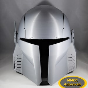 Puede incluir: Un casco gris impreso en 3D con una visera negra y una etiqueta MMCC Approved. El casco tiene un diseño futurista con detalles en los lados.