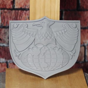 May include: A gray shield-shaped emblem featuring a detailed eagle with outstretched wings above a globe. The emblem is made of a solid material and has a raised relief design. The background is a mix of wood and brick.