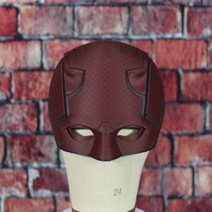 Könnte beinhalten: Eine rote Daredevil-Maske mit schwarzen Details und einer strukturierten Oberfläche. Die Maske hat zwei Augenlöcher und einen erhöhten Nasensteg.