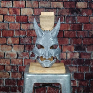 May include: A grey, detailed, 3D printed Oni mask with horns and a fierce expression. The mask is displayed on a wooden stand.