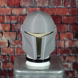 May include: A gray Mandalorian helmet, a replica of the iconic helmet from the Star Wars series. The helmet features a distinctive T-shaped visor and angular design. It is displayed on a white and black base, set against a brick wall background.