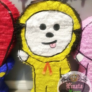 May include: A yellow and white pinata shaped like a cartoon character with a black outline. The character has a white face with black eyes, a black nose, and a red tongue sticking out. The pinata is decorated with orange and red streamers. The pinata is standing on a brown surface. The text "Pinata Kings" is visible in the lower right corner of the image.