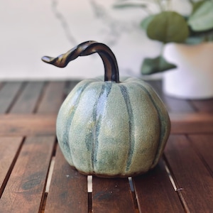 May include: A ceramic pumpkin with a green and tan finish, featuring vertical lines and a twisted brown stem. The decorative gourd sits on a wooden surface, with a blurred background.