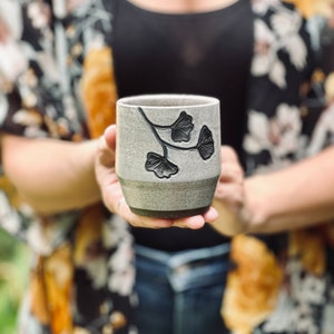 hands holding ceramic cup decorated with a speckled gray glaze and dark brown ginkgo carvings