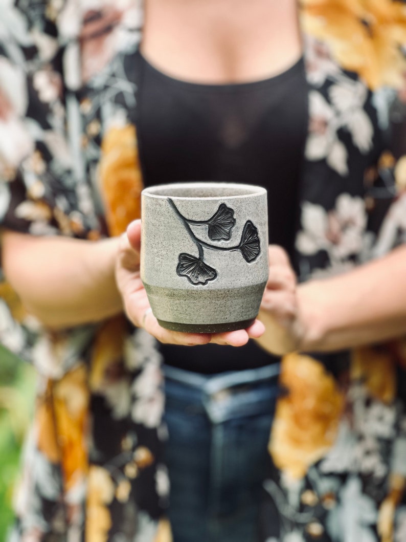 hands holding ceramic cup decorated with a speckled gray glaze and dark brown ginkgo carvings