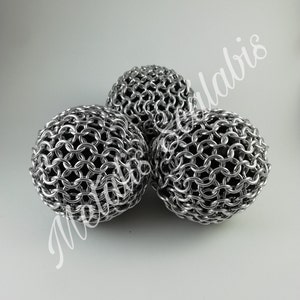 Op de afbeelding: Drie bolvormige decoratieve objecten gemaakt van verweven zilverkleurige metalen ringen. De bollen zijn dicht bij elkaar geplaatst, met een helder, reflecterend oppervlak. Het algehele effect is een moderne, industriële esthetiek. De objecten staan tegen een witte achtergrond.